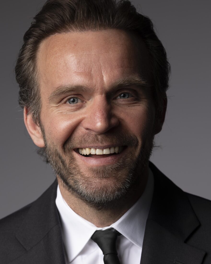 Jack Borkowski, smiling, in a dark suit and tie against a grey studio background.